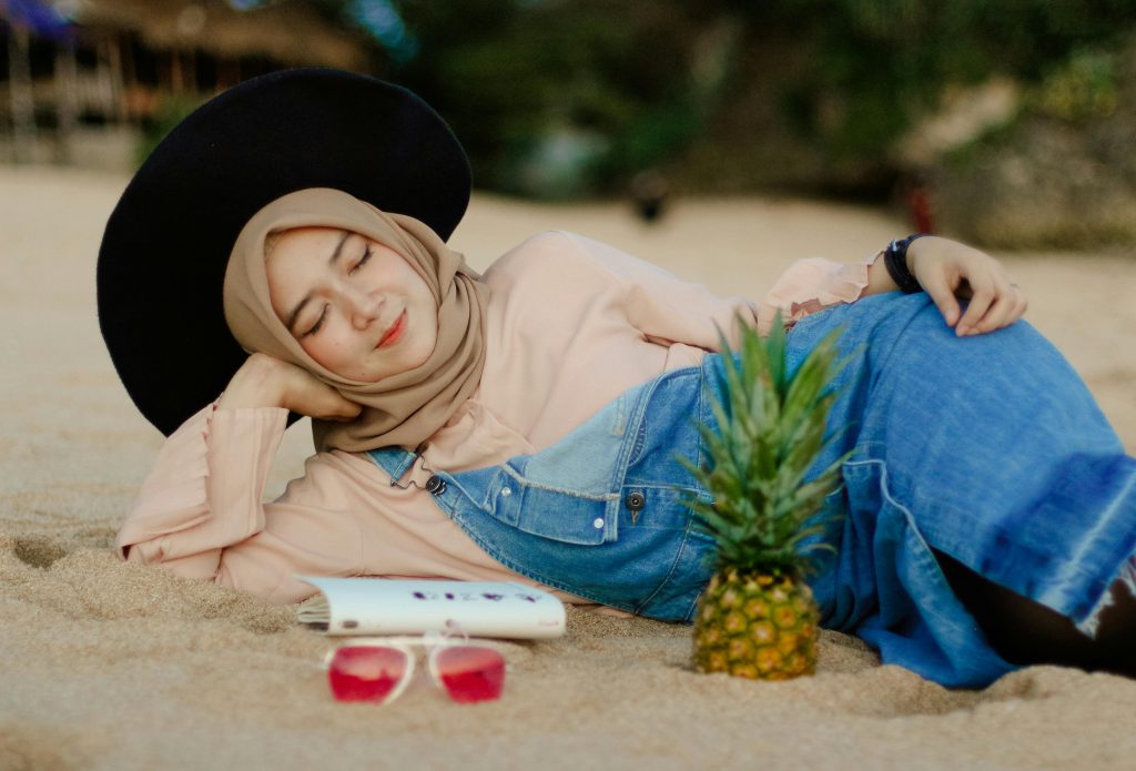 A young woman in a hijab relaxes on the beach near a pineapple, enjoying a sunny day in Jogja.