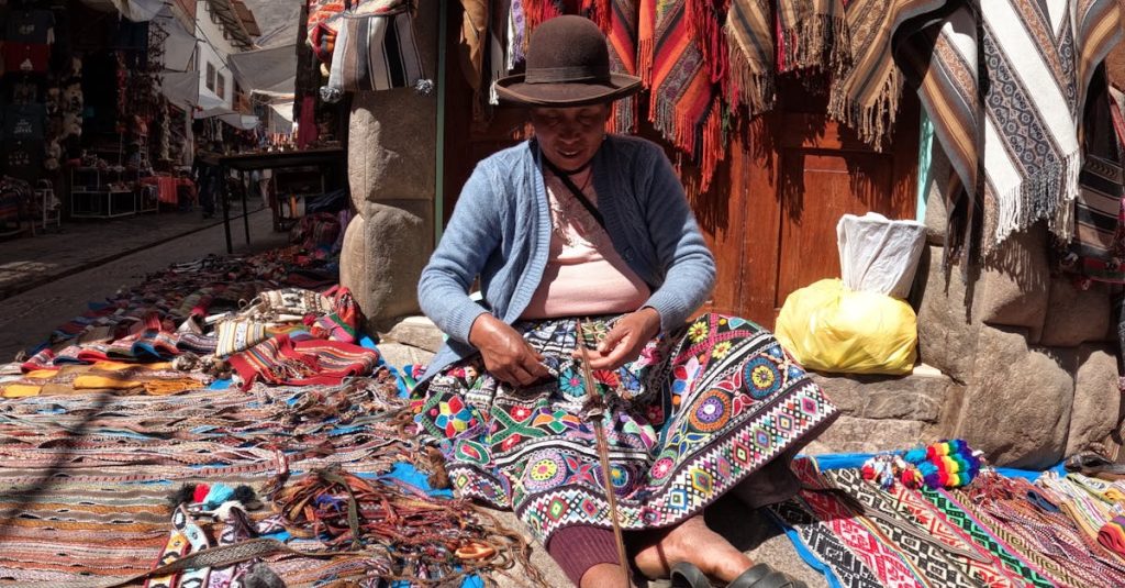 Woman weaving traditional textiles in a colorful outdoor market setting.
